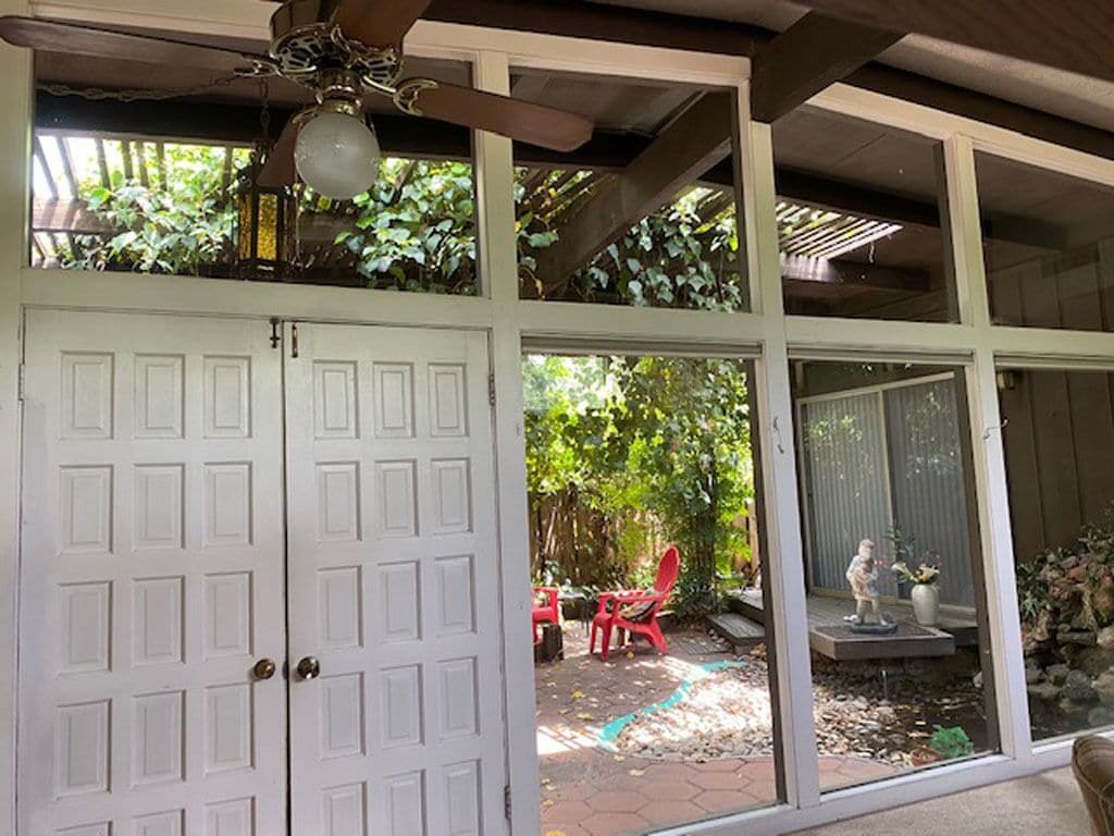 Un patio techado con ventilador de techo, una puerta blanca y vistas a un jardín con sillas rojas y una estatua.