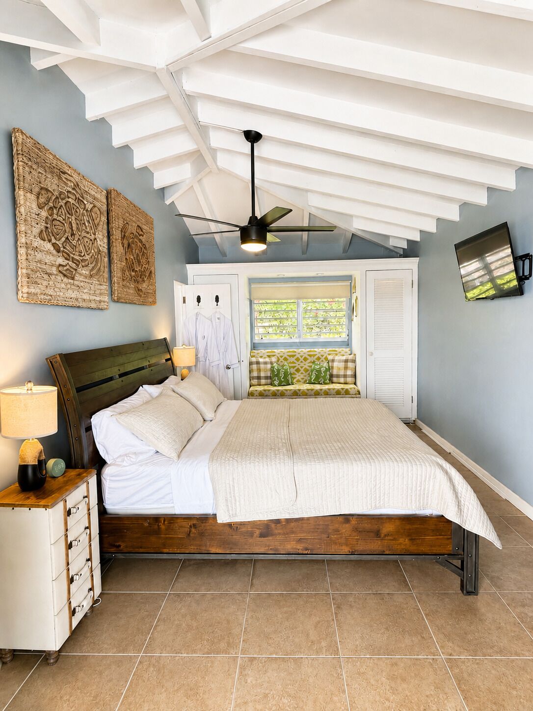 Un dormitorio con cama, mesitas de noche, ventilador de techo, obras de arte, una ventana con contraventanas y un televisor montado en la pared.
