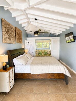 Une chambre à coucher comprenant un lit, des tables de chevet, un ventilateur de plafond, des œuvres d’art, une fenêtre avec des volets et un téléviseur fixé au mur.