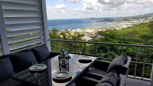 Un balcon avec une table et des chaises, donnant sur un paysage côtier avec des bâtiments et de la végétation.