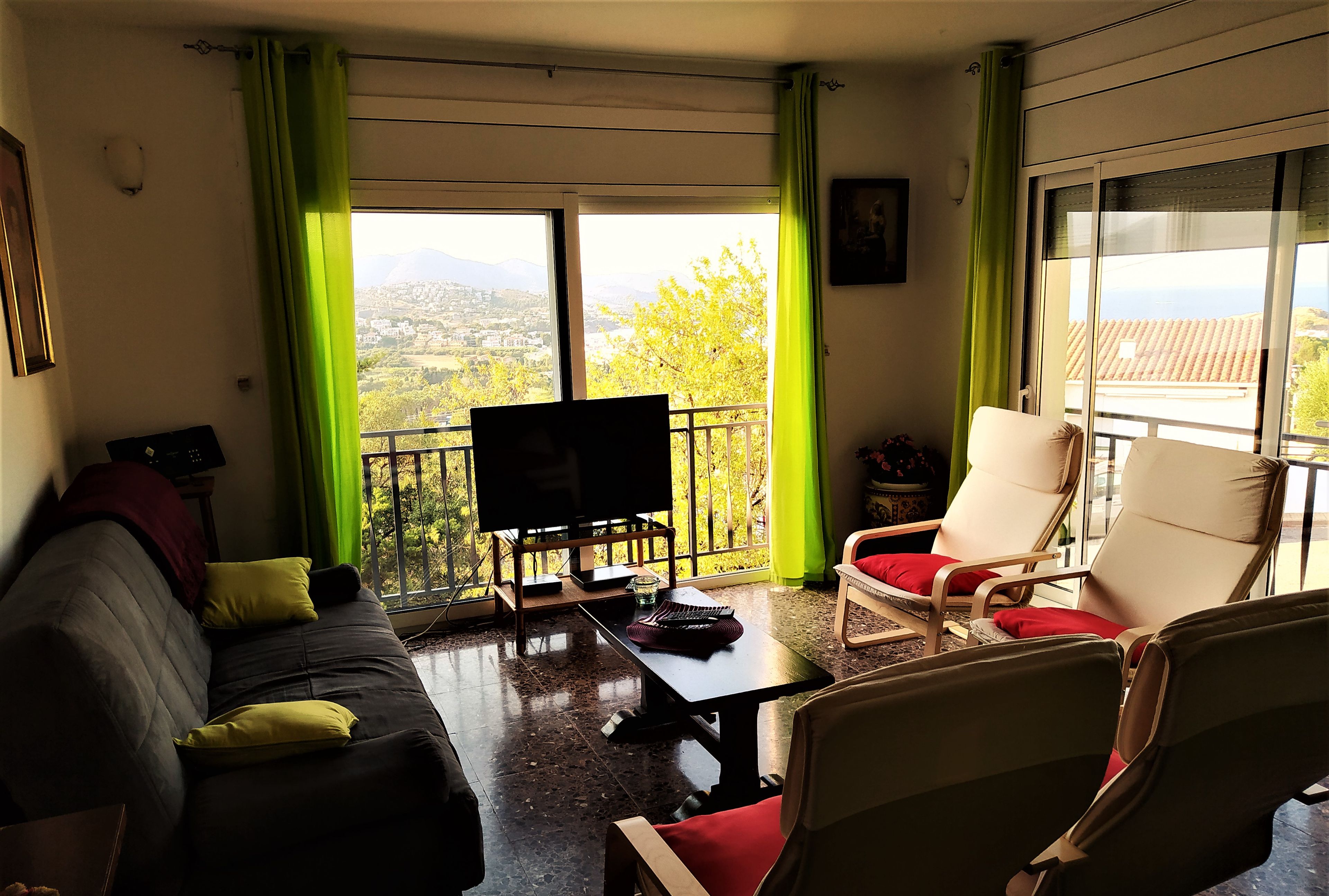 A living room with a sofa, a TV, and a table with a remote control. There is a balcony with a view of the city and mountains.