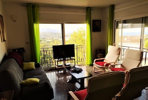 A living room with a sofa, a TV, and a table with a remote control. There is a balcony with a view of the city and mountains.