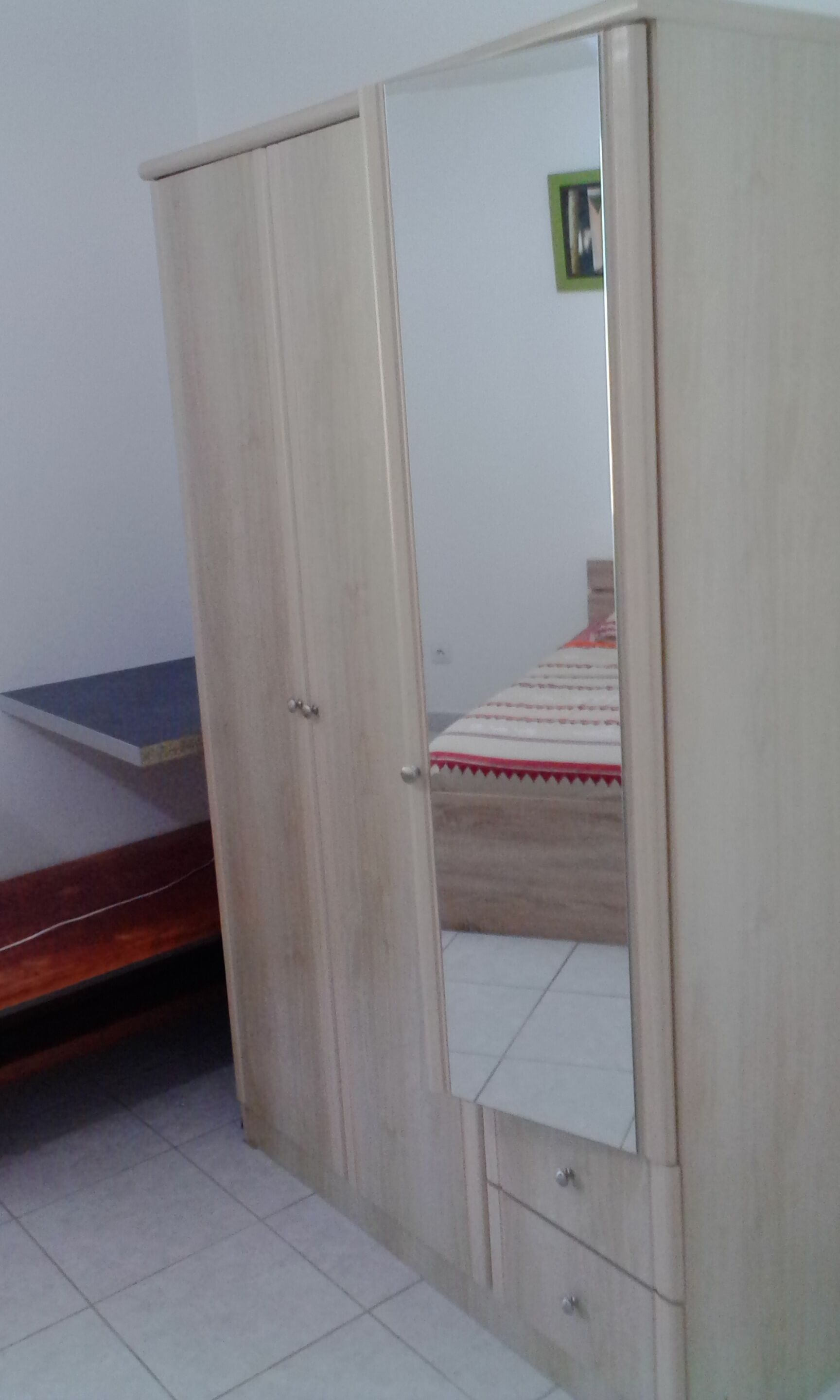 A room with a wooden wardrobe, a bed with a striped bedspread, and a small table.