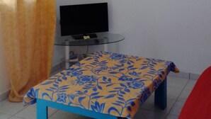 A room with a bed, a television, a table with a patterned tablecloth, a red chair, and a painting on the wall. - F2 air-conditioned with ocean view (Trois Rivieres)