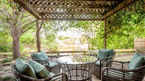 Un espace extérieur aménagé, abrité et doté de meubles en osier et d’une table en verre, offrant une vue sur un paysage naturel avec des arbres et une cascade.