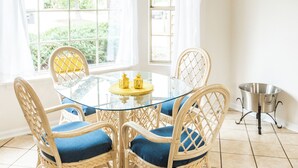 Un coin repas comprenant une table ronde en verre, quatre chaises en osier avec des coussins bleus, un ventilateur de plafond et une fenêtre agrémentée de rideaux blancs.