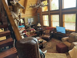A rustic cabin interior with a large deer head mounted on the wall, a cozy seating area with a leather sofa and armchairs, a television on a wooden table, and a large window with blinds.