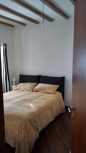 Un dormitorio con cama, vigas de madera en el techo, una mesita de noche con lámpara y una puerta.