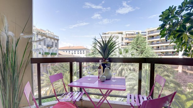 Un balcón con una mesa y sillas rosadas, una planta en maceta y vistas a edificios y árboles.