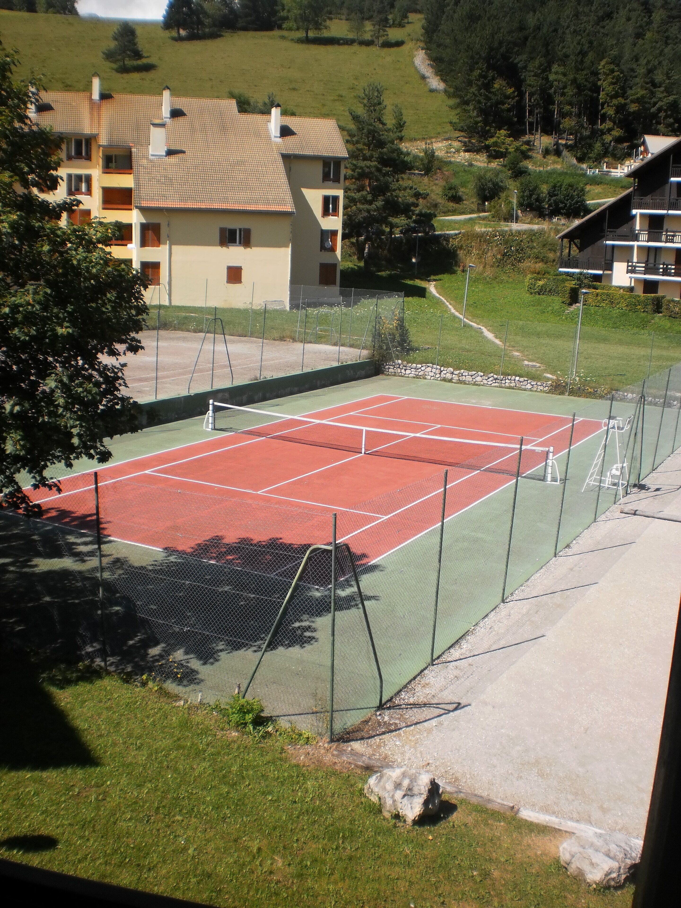 A tennis court with a net, surrounded by a green fence. There are buildings in the background.