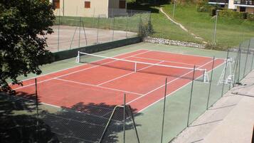 A tennis court with a net, surrounded by a green fence. There are buildings in the background.