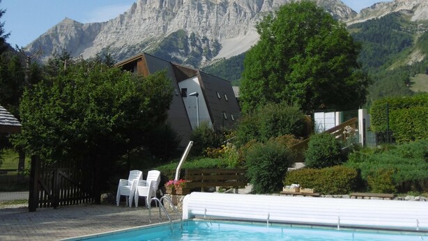 A swimming pool with a mountain backdrop, surrounded by greenery and a wooden deck with chairs.