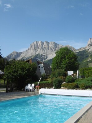 A swimming pool with a mountain backdrop, surrounded by greenery and a wooden deck with chairs.