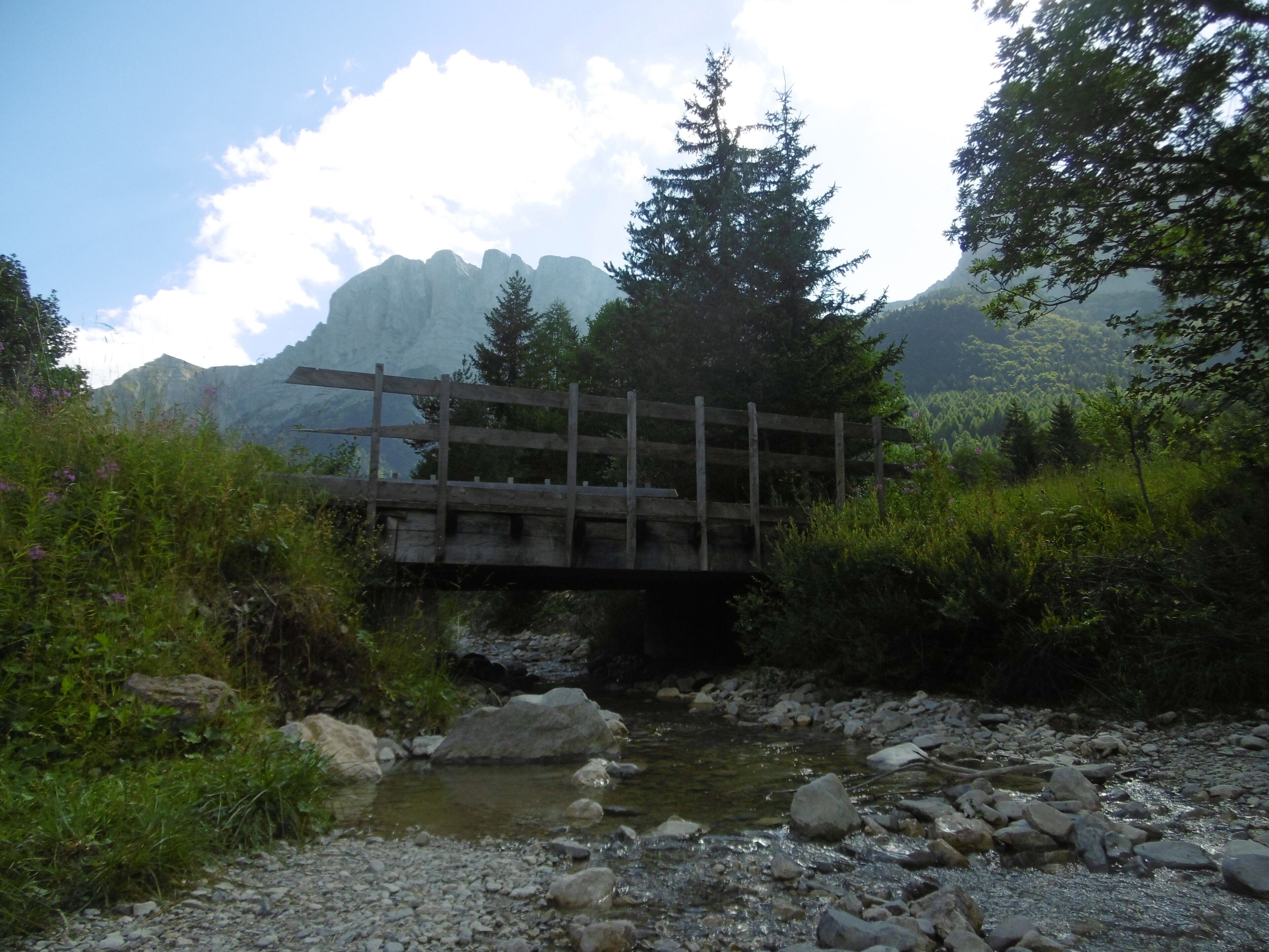 A wooden bridge over a stream with rocks and greenery on either side, set against a mountainous backdrop.