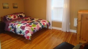 A bedroom with a wooden floor, a bed with a colorful floral bedspread, a window with white curtains, and a wooden dresser. - Great center of historic districts on Water Street (Charlottetown)