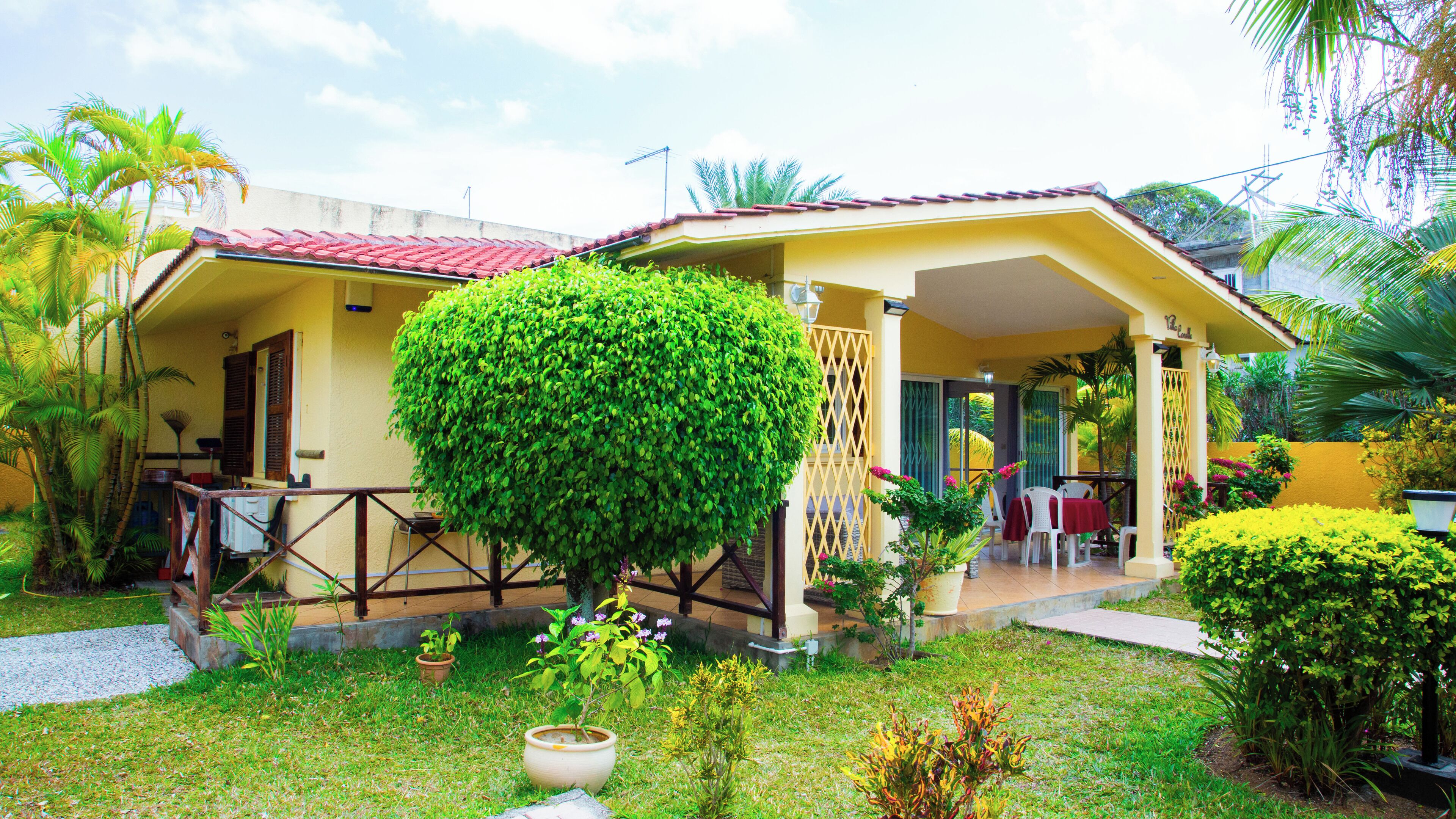 A yellow house with a red roof, a covered porch, and a well-maintained garden with various plants and a large bush.