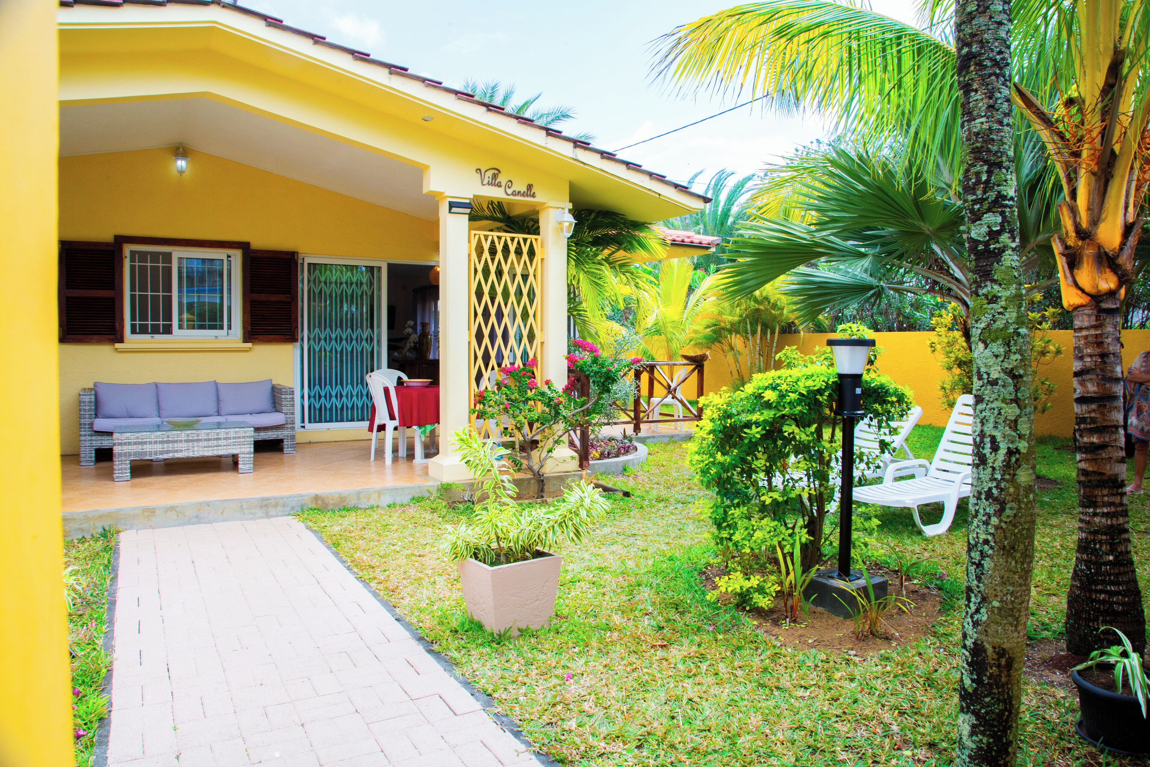 Una villa amarilla con un porche cubierto, mobiliario de exterior y abundante vegetación.