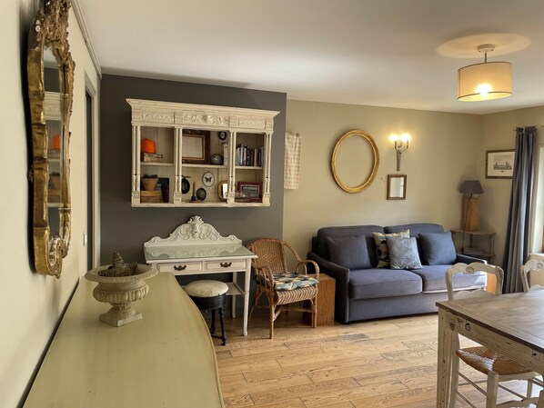 A living room with a grey sofa, a white decorative headboard, a wooden dining table, and a bookshelf.