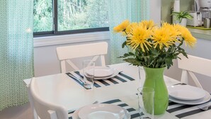Comedor con mesa dispuesta para cuatro, un jarrón de flores amarillas y una ventana con vista a follaje.