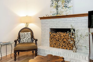A living room with a white brick fireplace, a wooden chair with a patterned cushion, a side table, and a lamp.