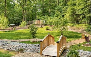 A wooden bridge over a stream in a forested area with a well-maintained lawn and a stone pathway.