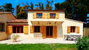 A two-story house with a tiled roof, wooden shutters, and a thatched roof over the entrance. There is a patio with outdoor furniture.