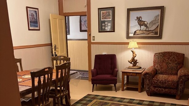 Un salon comprenant un coin repas, avec une table à manger en bois, des chaises, un fauteuil rouge, un tapis à motifs et des œuvres d’art encadrées accrochées aux murs.