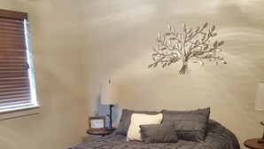 A bedroom with a bed, a wall clock, a decorative tree, and a small table with a lamp.