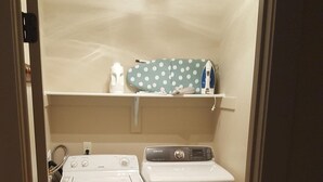 A compact laundry room with a front-loading washer and dryer, a small shelf with laundry supplies, and a trash bin.