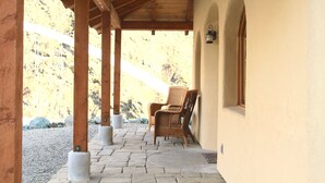 A covered outdoor area with stone flooring, wooden pillars, and a wicker chair. - Charming Casita With Spectacular River Views  (Riggins)