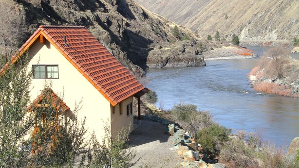A small house with a red-tiled roof near a river, surrounded by rocky hills. - Charming Casita With Spectacular River Views  (Riggins)