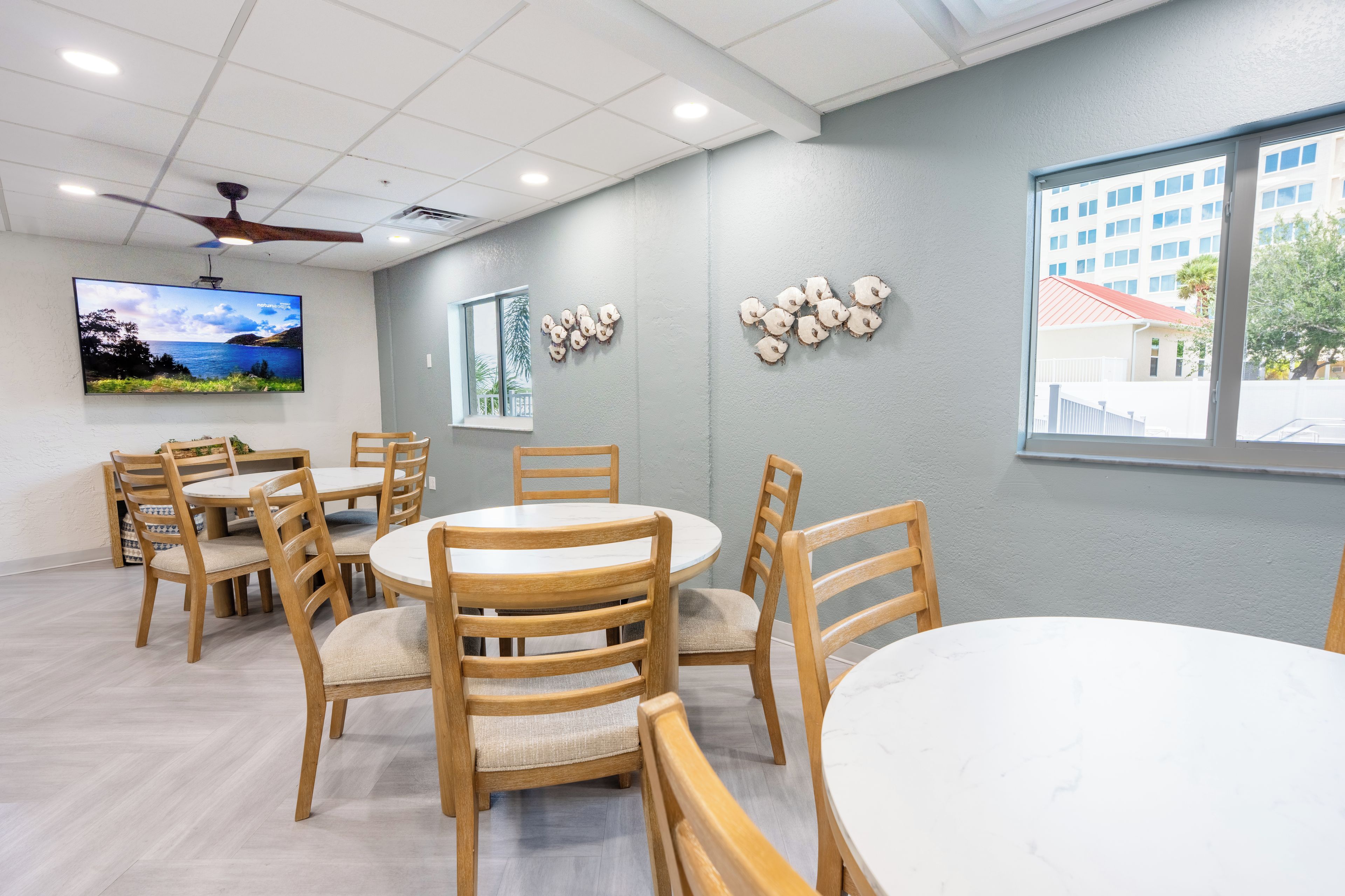 A conference room with round tables and wooden chairs, a flat-screen TV on the wall, and a window with a view of buildings and trees.