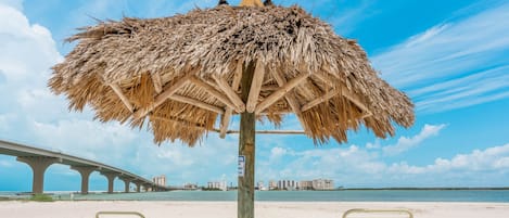 A beach with two metal lounge chairs under a thatched umbrella, a wooden post with a sign, and a bridge in the background.