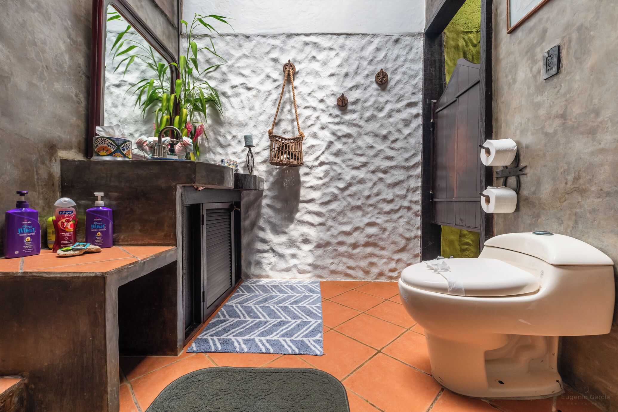 Ein kleines Badezimmer im rustikalen Stil, mit einer weißen Toilette, einem Waschbecken mit Toilettenartikeln, einer dekorativen Wand und einem Fenster mit Blick.