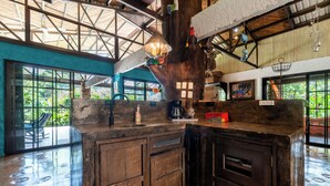 A rustic kitchen with wooden cabinets and a large island, a hanging light fixture, and a view of the outdoors through large windows. - Breakfast With Toucans And Monkeys (San Miguel)
