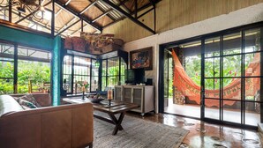 A spacious living area with a sofa, a coffee table, and a television. Large windows offer views of greenery outside. - Breakfast With Toucans And Monkeys (San Miguel)