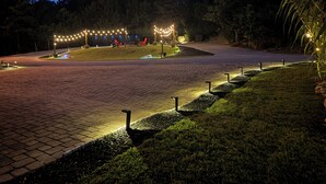 Un chemin pavé mène à un espace aménagé avec des chaises et une table, le tout entouré d’arbres et d’une pelouse.