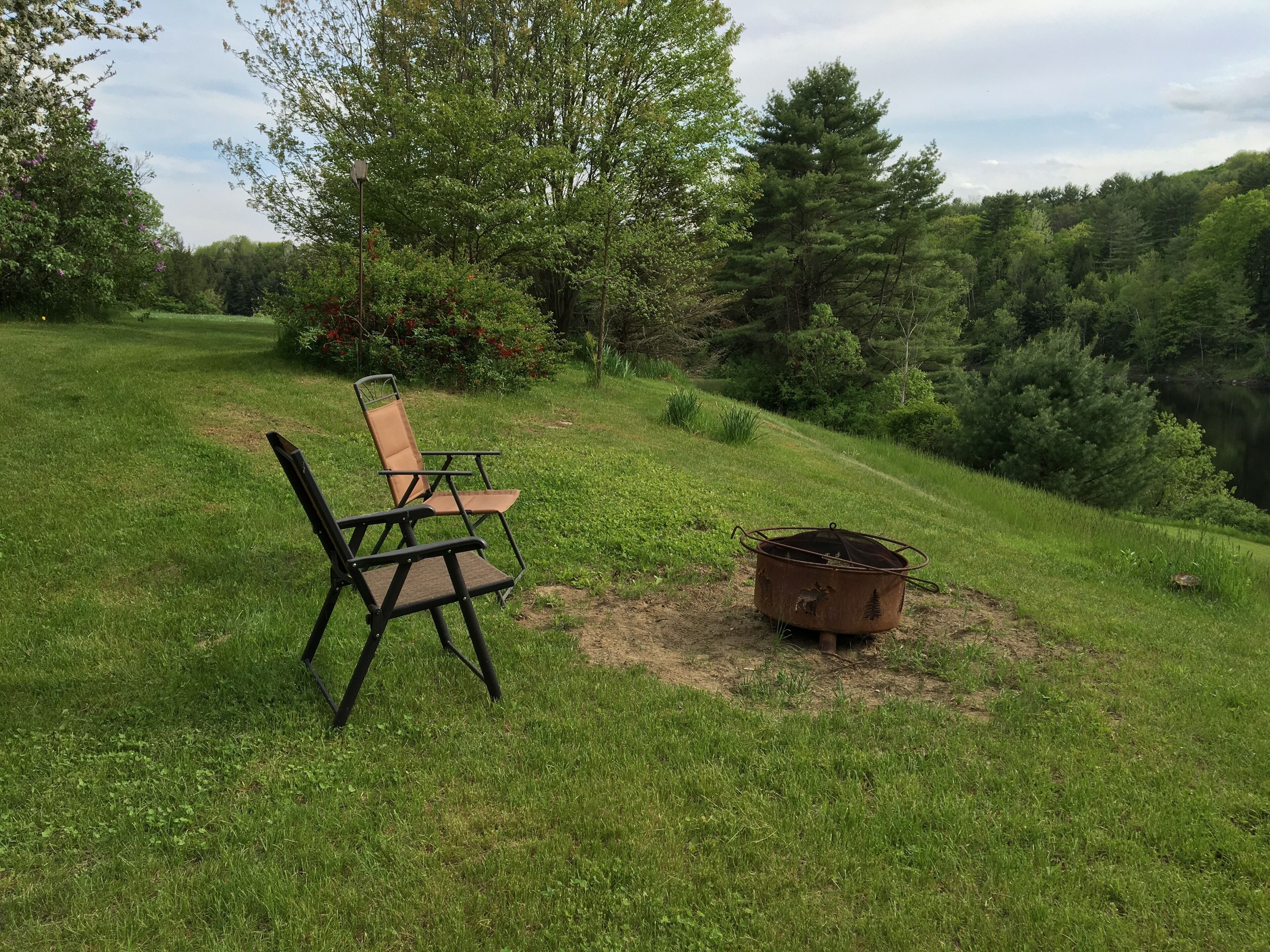 Un espace extérieur verdoyant avec un foyer en métal et deux chaises.