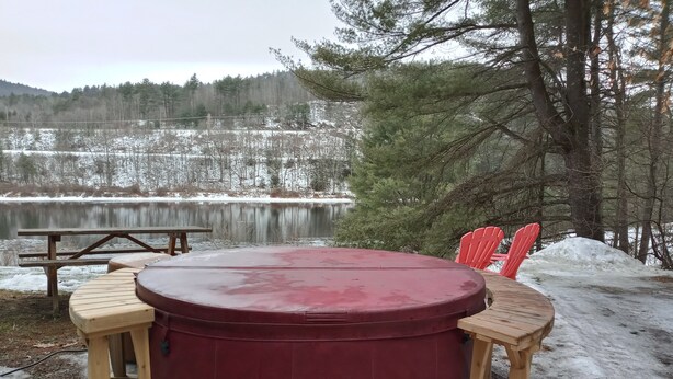 Un bain à remous circulaire en bois, surmonté d’un couvercle rouge, entouré de chaises en bois, installé sur un sol enneigé, avec vue sur un lac et des arbres en arrière-plan.