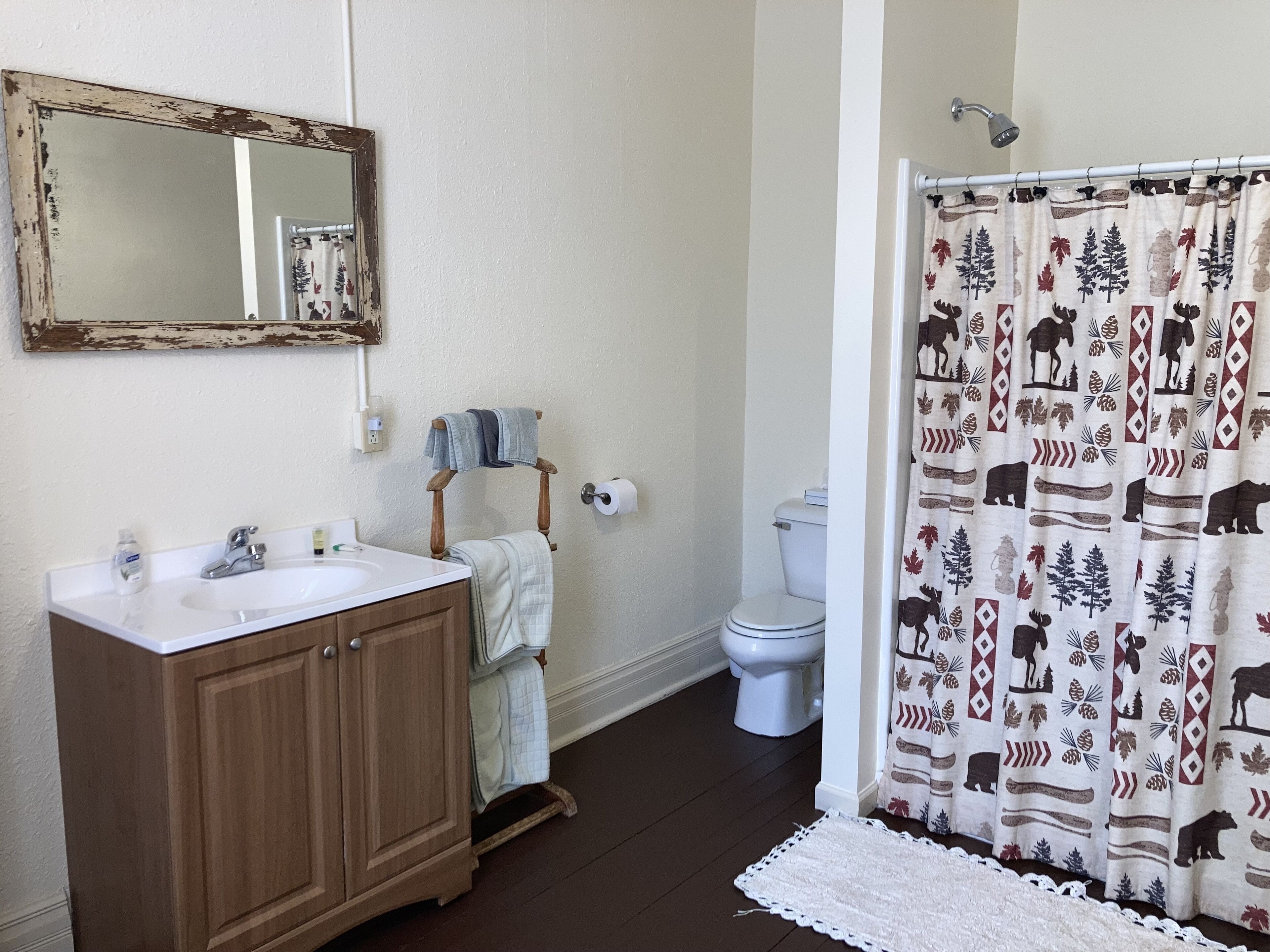 A bathroom with a wooden vanity, a white sink, a mirror, a towel rack, a toilet, and a shower curtain with a forest and animal print.