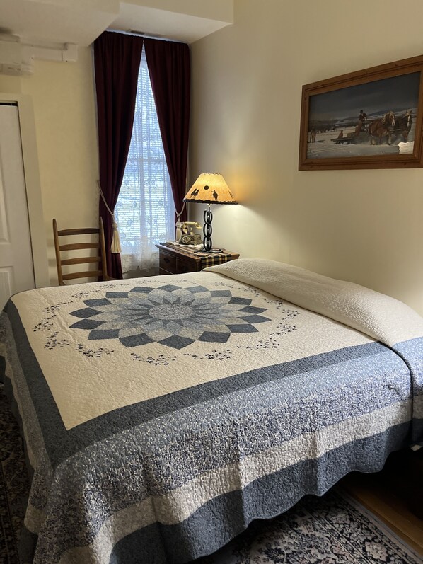 Un dormitorio con una cama grande, una mesita de noche con lámpara, una silla, un ventanal con cortinas y un cuadro en la pared.