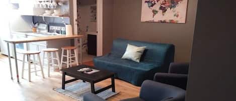 Sala de estar con sofá, una mesa de centro y dos sillas. Cuenta con una zona de cocina con taburete y mostrador. En la pared, se exhibe un mapa del mundo.