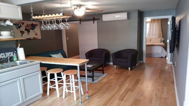 A compact kitchen with a wooden countertop and stools, a dark sofa, and two armchairs in a living area with hardwood flooring. - Wine Alley West Suite (Hermann)