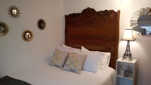 A neatly made bed with floral pillows, a wooden headboard, and a bedside table with a lamp and a mirror. - The House on Wine Alley (Hermann)