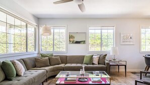 A living room with a sectional sofa, a glass coffee table, and large windows with a view of greenery. - Movie Colony Home with Private Casita, Pool & Spa! (Palm Springs)