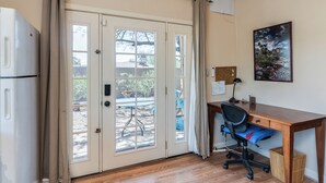 Una stanza con una scrivania in legno, una sedia, un frigorifero e una porta scorrevole in vetro che dà su uno spazio esterno.