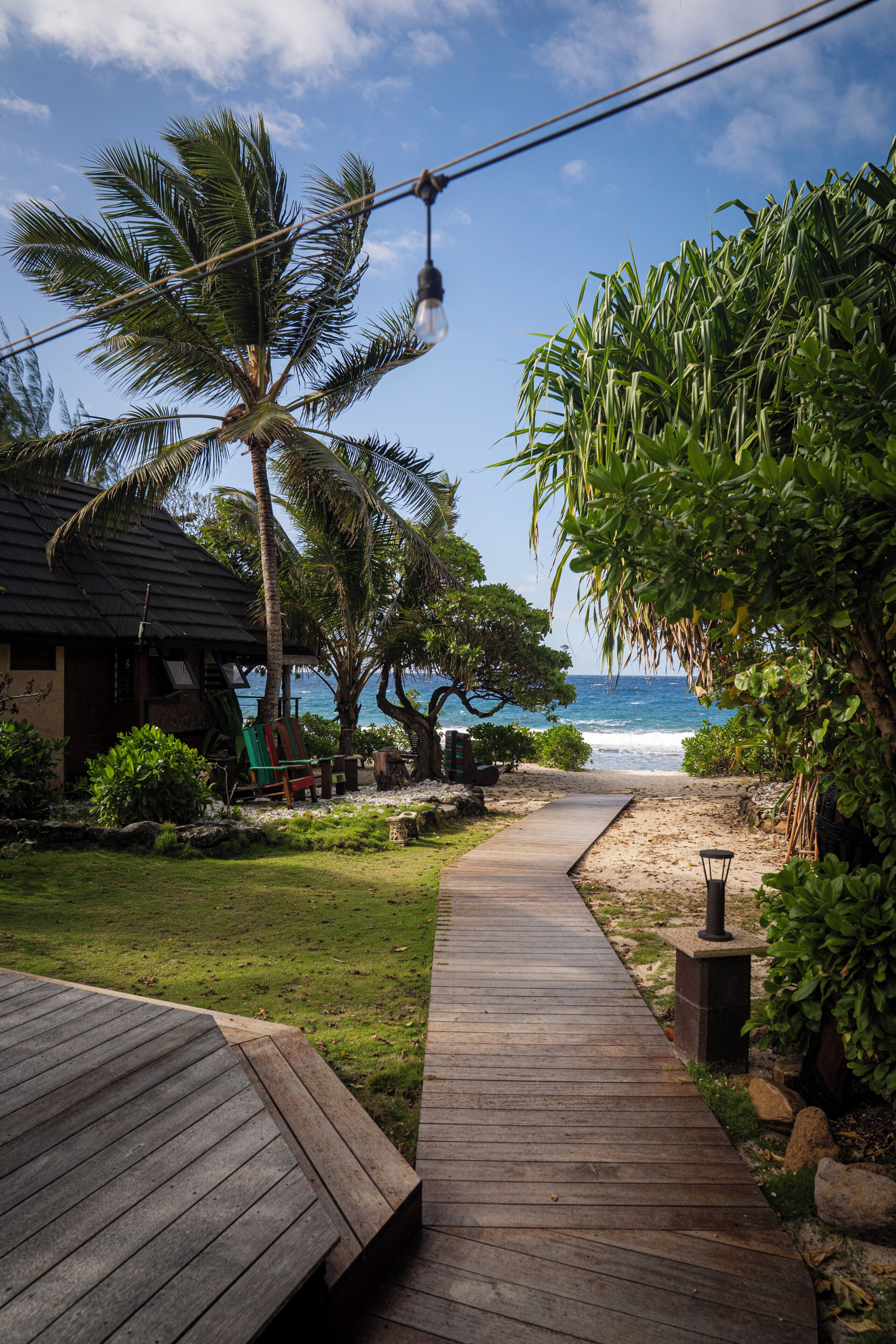 A wooden pathway leading to a beach with palm trees and a building with a dark roof.