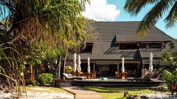 A resort with a wooden walkway leading to a building with a dark roof, surrounded by palm trees and outdoor seating. - Green Lodge Moorea (Moorea-Maiao)