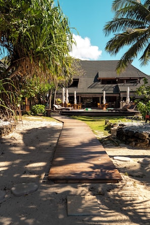 A resort with a wooden walkway leading to a building with a dark roof, surrounded by palm trees and outdoor seating. - Green Lodge Moorea (Moorea-Maiao)