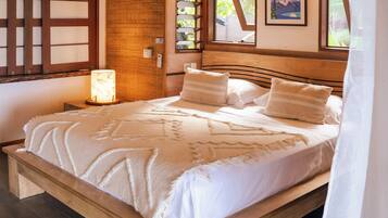 A wooden-framed bed with a white bedspread and pillows, a wall-mounted air conditioner, and a window with a view of greenery.